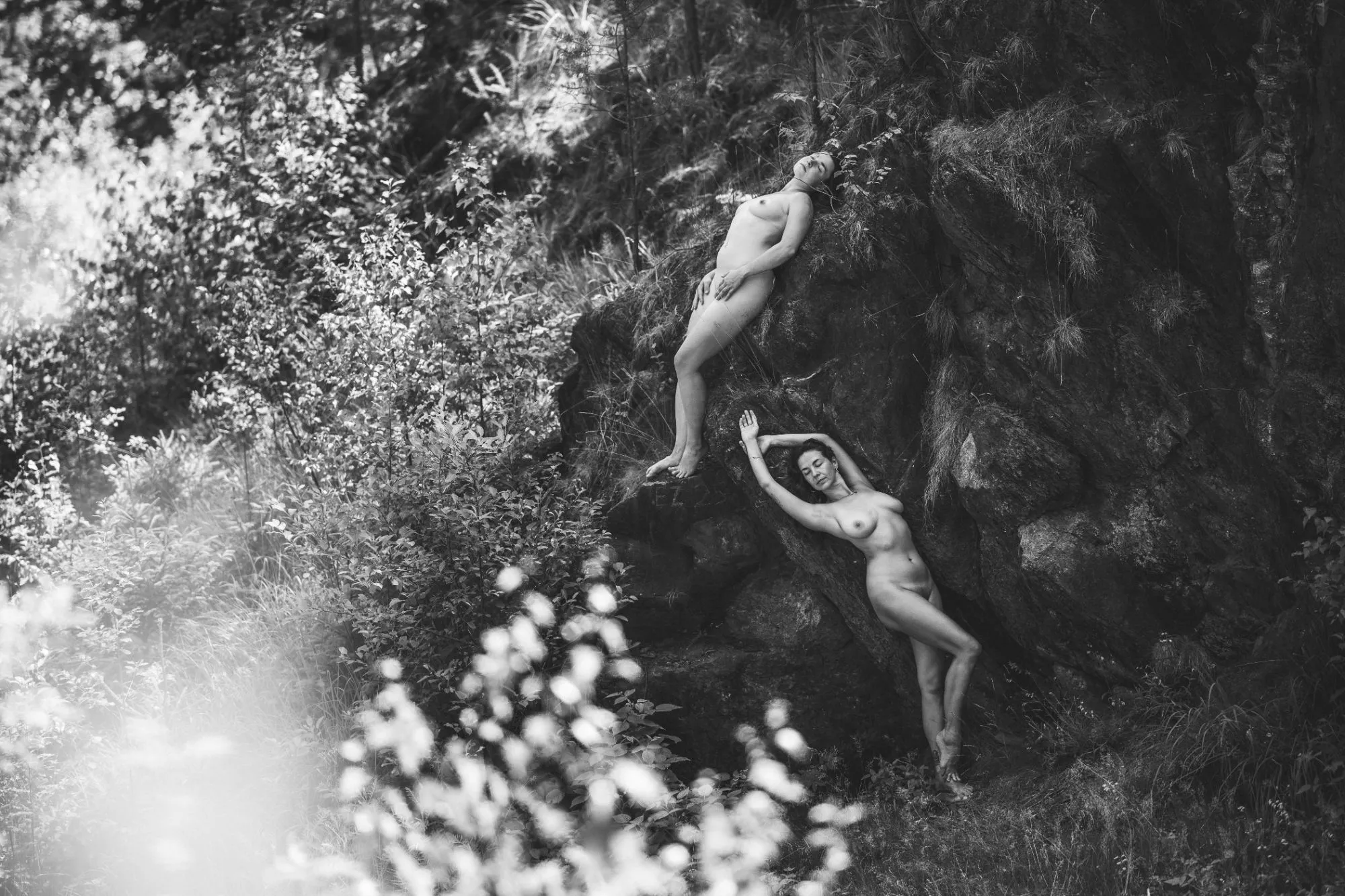 Zwei nackte Frauen lehnen an einer bewachsenen Felswand im Wald, links helles Laub im Gegenlicht