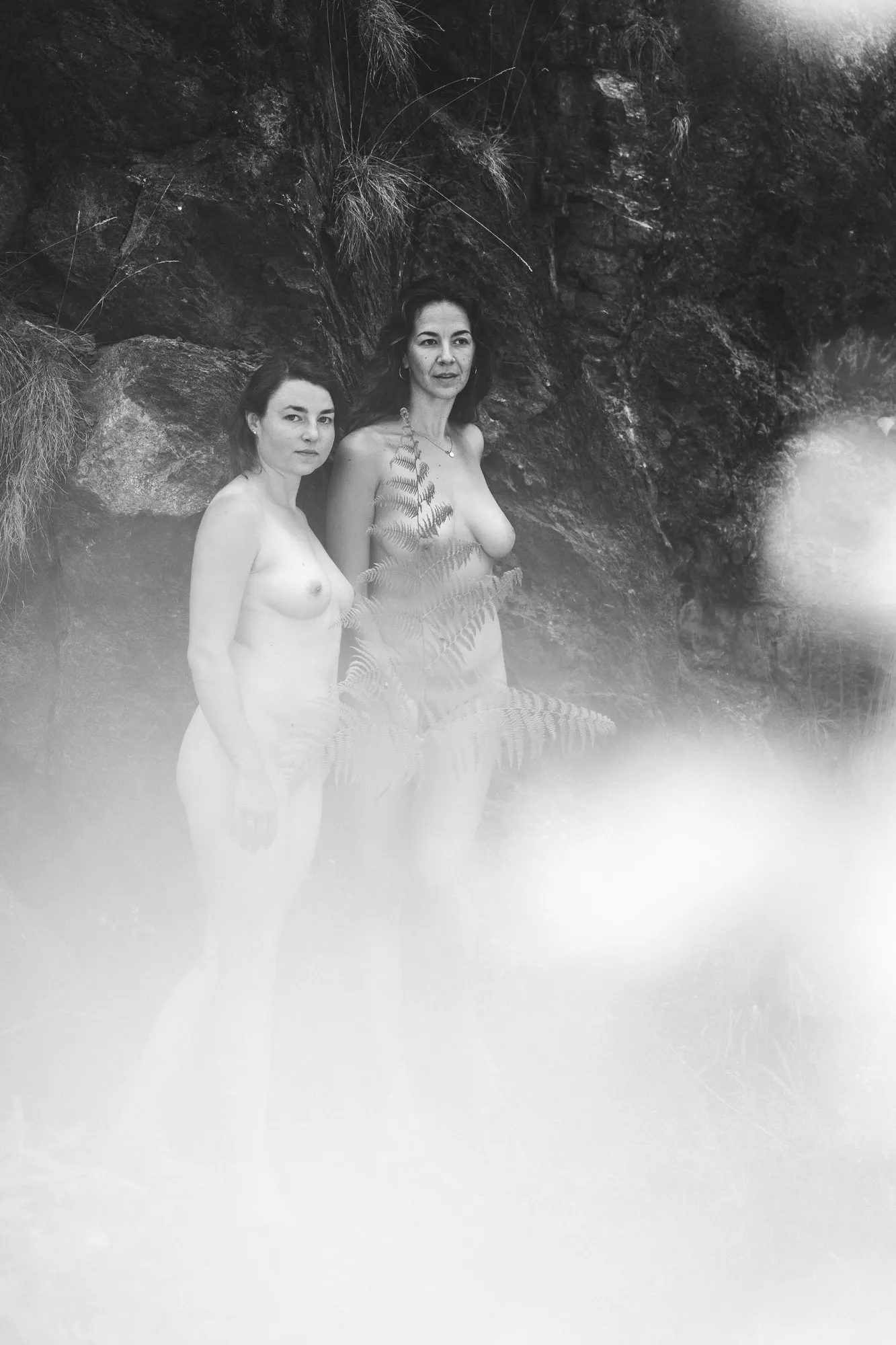 Zwei nackte Frauen stehen vor einer dunklen Felswand, ein Farnwedel vor ihren Körpern, im Vordergrund ein heller Lichtschleier